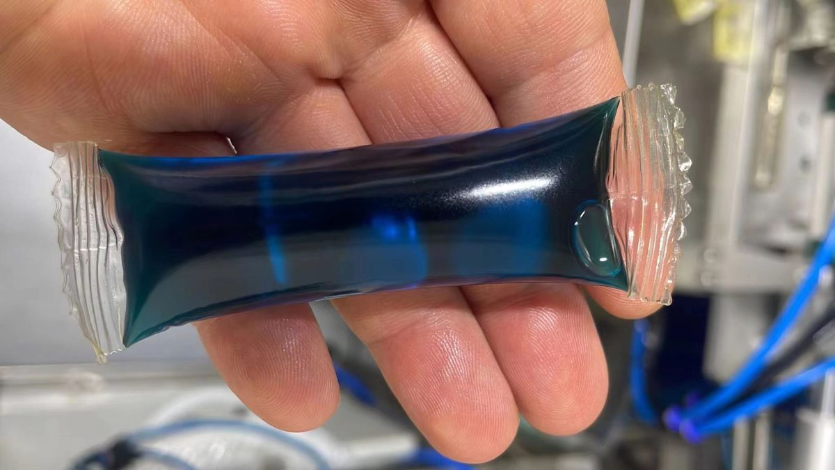 A filled and sealed water-soluble liquid pack held in front of a filling machine